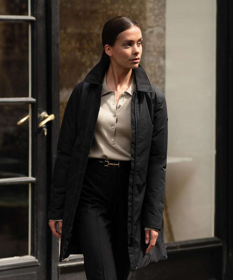 A person with dark hair pulled back is wearing the Nimbus Womens Folcroft jacket, a beige collared shirt, and black trousers while standing near a glass door with a brass handle.