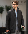 A man wearing glasses, a light blue dress shirt, grey suit trousers, and the Nimbus Folcroft jacket stands outdoors with a black bag and dark umbrella. Large plants and a blurred building are visible in the background.