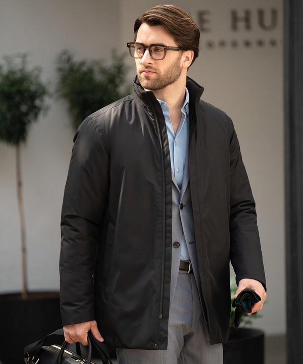 A man wearing glasses, a light blue dress shirt, grey suit trousers, and the Nimbus Folcroft jacket stands outdoors with a black bag and dark umbrella. Large plants and a blurred building are visible in the background.