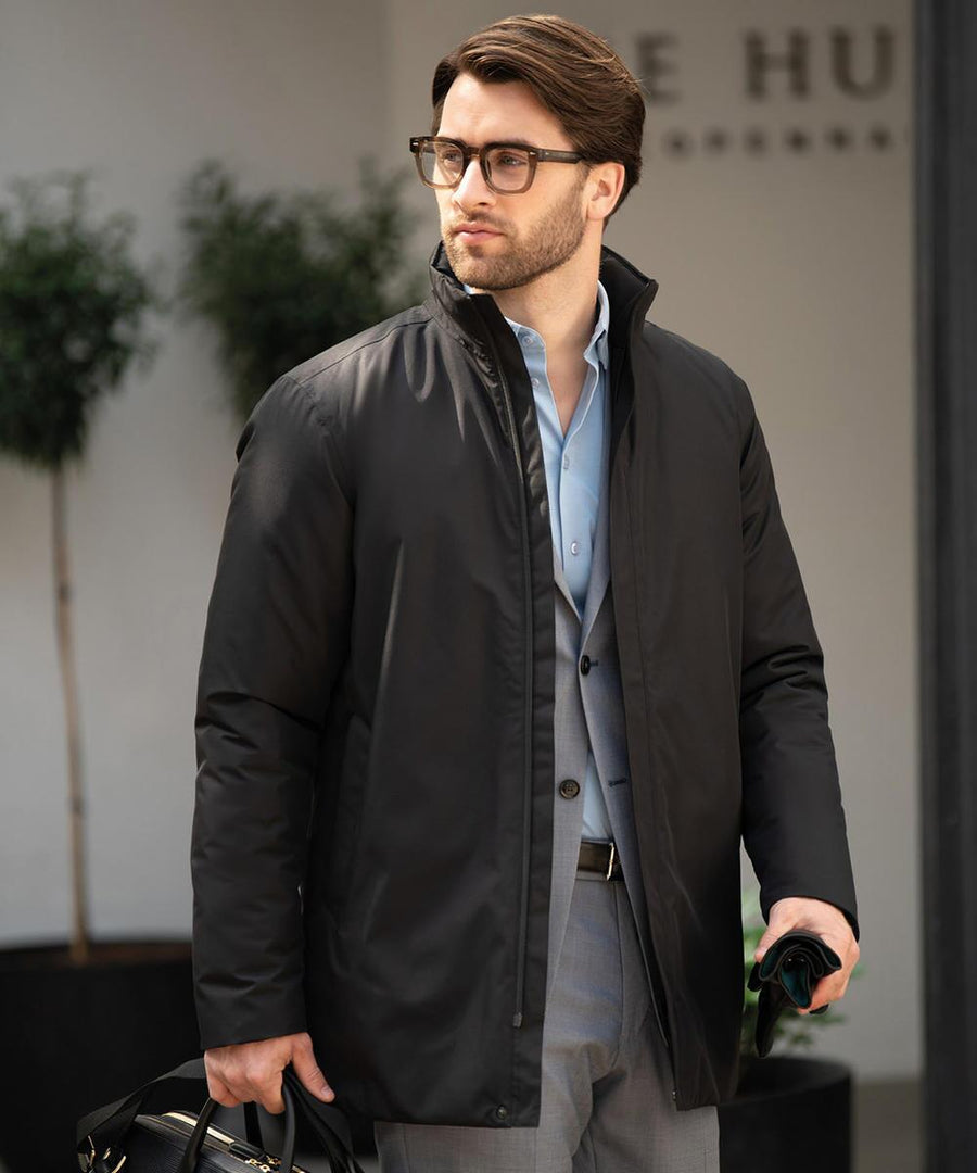A man wearing glasses, a light blue dress shirt, grey suit trousers, and the Nimbus Folcroft jacket stands outdoors with a black bag and dark umbrella. Large plants and a blurred building are visible in the background.