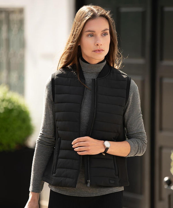 A woman with long brown hair stands outdoors near a dark door and greenery, wearing the Nimbus Women's Vesper gilet over a grey polo neck jumper and adjusting its zip.