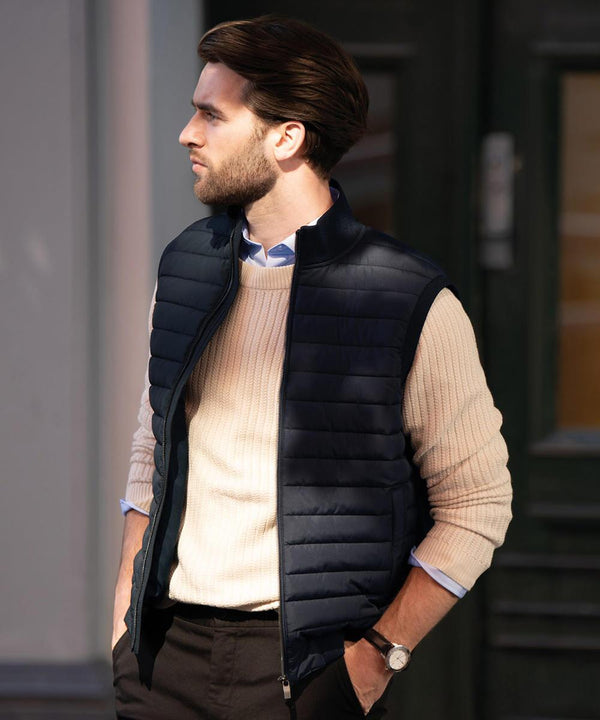 A man with brown hair stands outdoors, wearing the Nimbus Vesper gilet over a light beige jumper and dark trousers. He has his hands in his pockets and looks to the left with a neutral expression.