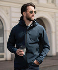 A man in sunglasses and a Nimbus Yosemite jacket holds a takeaway coffee cup while standing outdoors in front of a building with arched windows.