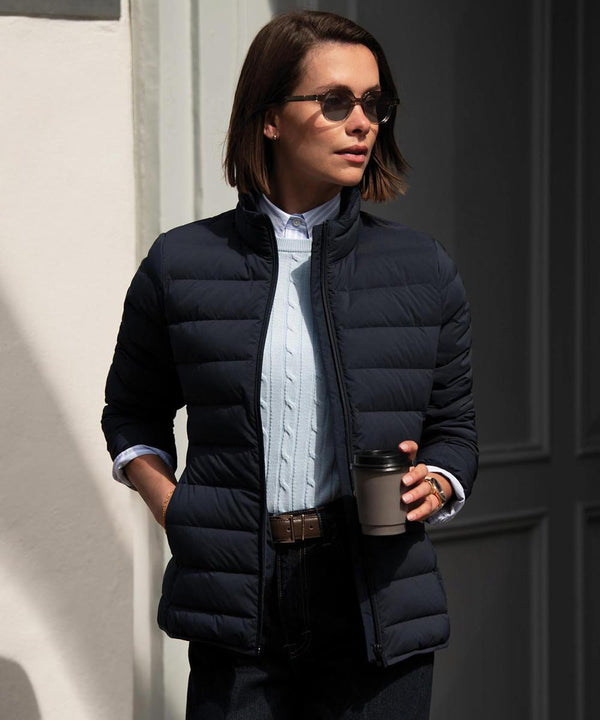 A woman with short brown hair stands near a doorway, wearing the Nimbus Women's Avalon jacket, sunglasses, and holding a reusable coffee cup. She layers the navy puffer jacket over a light blue jumper and collared shirt.