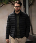 A man with brown hair and a beard is wearing black-rimmed glasses, an olive green jumper, beige trousers, and the Nimbus Avalon jacket. He is outdoors, holding a brown bag and looking slightly to the side.