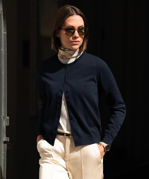 A woman in the Nimbus Women's Brighton cardigan stands outdoors, styled with sunglasses, a white polo neck, cream trousers, and a patterned scarf, one hand in her pocket as she looks to the side.