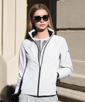 A woman with straight brown hair and dark sunglasses stands outdoors, leaning against a beige building and wearing the Nimbus Women’s Prescott jacket—showcasing comfortable, streamlined elegance with one hand in her pocket.