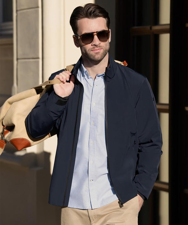 A man outdoors carries a beige bag with brown straps while wearing Nimbus’s Prescott—an outfit of comfortable, streamlined elegance—featuring sunglasses, a navy jacket, light blue button-up shirt, and beige trousers.