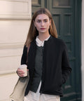 A person with straight, light brown hair and neutral expression stands outside holding a coffee cup and beige tote, wearing the Nimbus Womens Westlake performance hybrid jacket over a white shirt and grey cardigan. Green door, white wall behind.