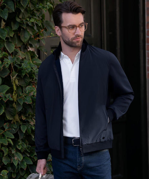 A man with short brown hair, a beard, and glasses stands outdoors by a green leafy wall, wearing the Nimbus Westlake performance hybrid jacket over a white polo and blue jeans. He holds an item in his left hand and looks to his right.