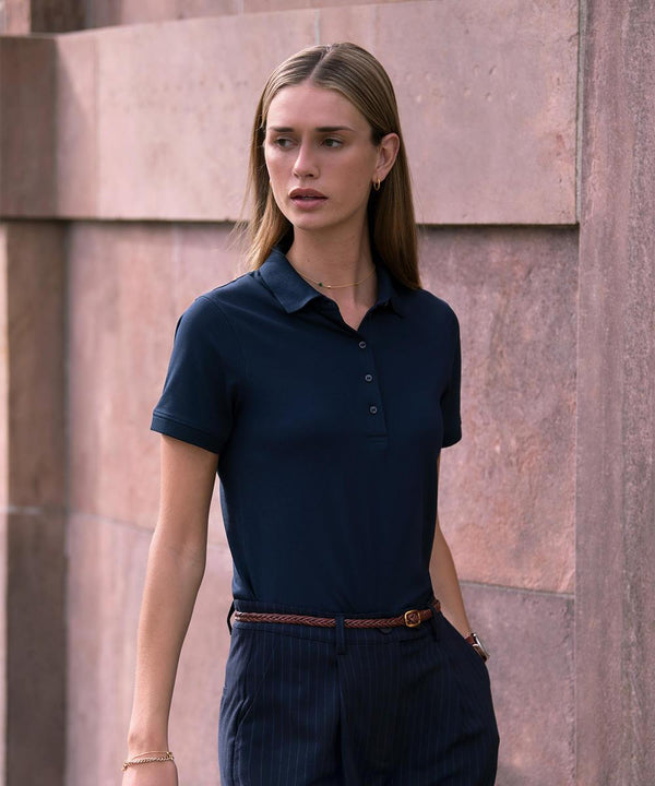 A person with long, straight hair wears the Nimbus Womens Altavista tri-blend deluxe polo and pinstriped trousers, standing against a textured stone wall and looking to the side with a neutral expression.