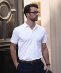 A man in glasses and dark trousers stands indoors near a dark decorative door, wearing a Nimbus Altavista tri-blend deluxe polo. He holds a green item in his left hand and looks off to the side with a neutral expression.