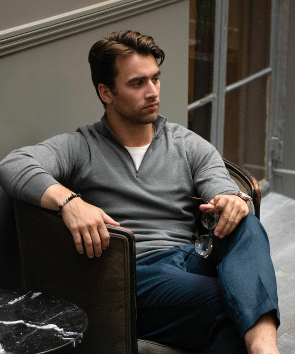 A man with dark hair, wearing a gray long-sleeve shirt and blue pants, sits on a chair indoors, holding a pair of Nimbus NB121 glasses while looking to his left.