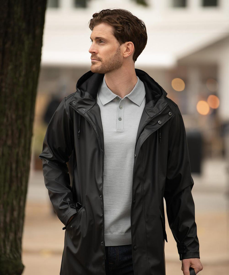 A man with short brown hair and a trimmed beard stands outdoors, wearing the Nimbus York unisex raincoat in black over a light grey polo shirt. He looks to the side with one hand in his coat pocket.