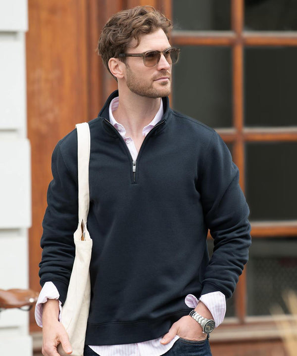 A man with short brown hair and sunglasses wears the Nimbus Glendale unisex 1/4-zip sweatshirt in navy over a light pink collared shirt, holding a beige canvas tote bag whilst standing by a window with wooden frames.