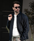 A man stands outdoors with a bag strap over his shoulder, wearing the Nimbus Detroit unisex jacket and sunglasses. A plant and a dark door are visible in the background.