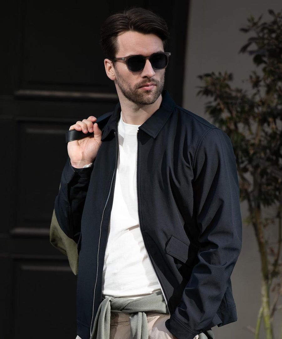 A man stands outdoors with a bag strap over his shoulder, wearing the Nimbus Detroit unisex jacket and sunglasses. A plant and a dark door are visible in the background.