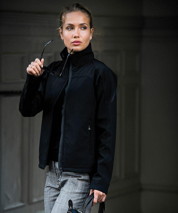 A person wearing a black jacket and gray pants holds NB30F sunglasses and a Nimbus bag, standing in front of a dark background.