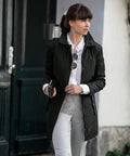 A woman with brown hair in a ponytail wears a black coat over a white shirt and gray pants, holding a Nimbus NB40F and sunglasses, walking outdoors near a building entrance.