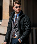 A bearded man wearing sunglasses, a black coat, a blazer, a tie, and an NB40M by Nimbus stands outside a building.