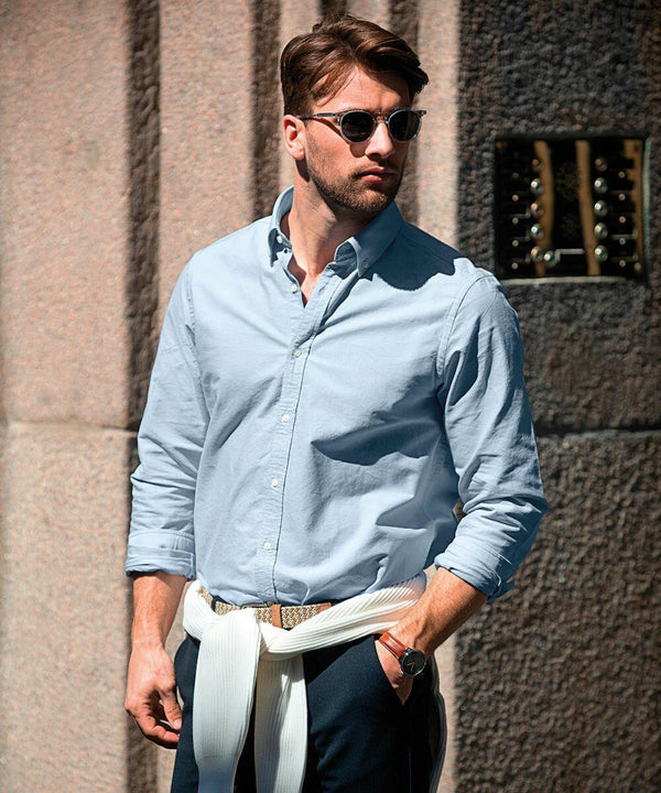 Man wearing Nimbus NB45M sunglasses and a light blue shirt stands against a stone wall with his hands in his pockets and a white sweater tied around his waist.