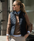 Woman wearing Nimbus NB46F sunglasses, a black quilted vest, beige sweater, and light pants standing outdoors in sunlight.