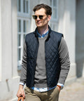 A man wearing a grey sweater, blue quilted vest, and Nimbus NB46M sunglasses is walking outside near a building.