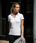 A woman wearing a white polo shirt and black pants is standing outdoors, holding a Nimbus NB52F, and looking to the side.