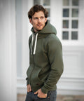 A person wearing a Nimbus NB55M green hoodie with white drawstrings and jeans stands outdoors in front of a building with large windows.