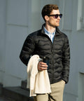 Man in sunglasses wearing a black puffer jacket, holding a white Nimbus NB60M sweater and a travel mug, walking outside.