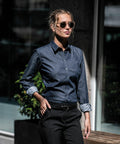 A woman wearing Nimbus NB65F sunglasses and a denim button-down shirt stands outdoors. She has her hands in her pockets and is in front of a blurred background with plants and glass windows.