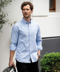 A man with wavy brown hair and a beard wears a light blue button-up shirt with rolled-up sleeves. He is standing outdoors holding a Nimbus NB66M jacket in his left hand.