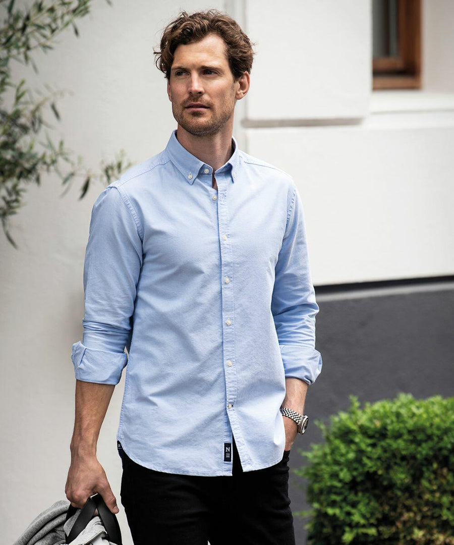 A man with wavy brown hair and a beard wears a light blue button-up shirt with rolled-up sleeves. He is standing outdoors holding a Nimbus NB66M jacket in his left hand.