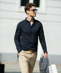 Man wearing Nimbus NB71M sunglasses, a navy long-sleeve shirt, and beige pants, holding a jacket and a shopping bag, walking outside near a white building.