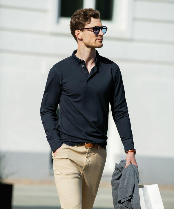 Man wearing Nimbus NB71M sunglasses, a navy long-sleeve shirt, and beige pants, holding a jacket and a shopping bag, walking outside near a white building.