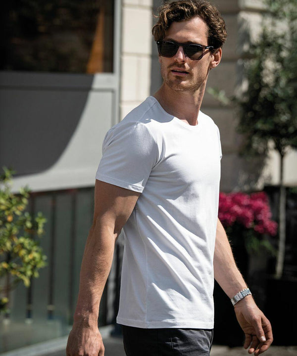 A man wearing Nimbus NB73M sunglasses and a white t-shirt walks outdoors, with plants and a building visible in the background.