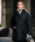 A person wearing a black winter coat stands outdoors, holding an umbrella and a checkered cloth bag. They appear to be in an urban setting, with buildings blurred in the background. The umbrella they are holding is the NB77F by Nimbus.