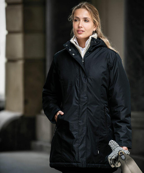A person wearing a black winter coat stands outdoors, holding an umbrella and a checkered cloth bag. They appear to be in an urban setting, with buildings blurred in the background. The umbrella they are holding is the NB77F by Nimbus.