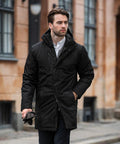 A man with brown hair, wearing a black coat and white shirt, holds Nimbus NB77M gloves and walks on a street with beige buildings in the background.