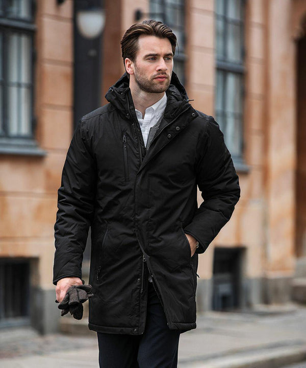A man with brown hair, wearing a black coat and white shirt, holds Nimbus NB77M gloves and walks on a street with beige buildings in the background.