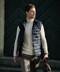 A man stands outdoors wearing a cream turtleneck, black puffer vest, and brown pants, holding a Nimbus NB79M.