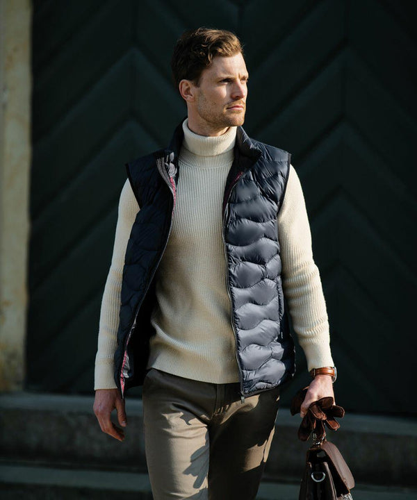 A man stands outdoors wearing a cream turtleneck, black puffer vest, and brown pants, holding a Nimbus NB79M.
