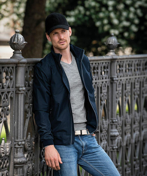 A person wearing a black NB82M jacket and a Nimbus cap leans against a decorative metal fence in an outdoor setting.