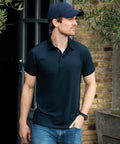 A man wearing a Nimbus NB86M polo shirt and cap stands outdoors in front of a building with a brick wall, looking to his left.
