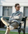 A man with sunglasses, wearing a Nimbus NB87M and khaki pants, sits casually on a bench with his legs crossed and arms folded.