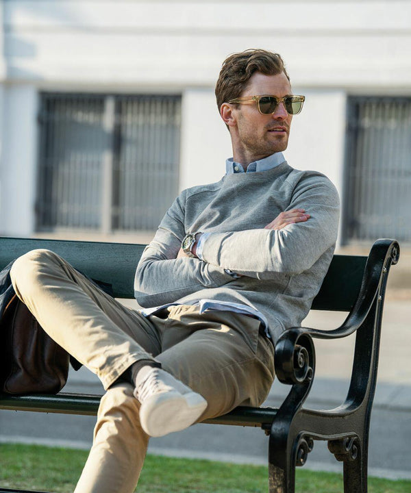 A man with sunglasses, wearing a Nimbus NB87M and khaki pants, sits casually on a bench with his legs crossed and arms folded.