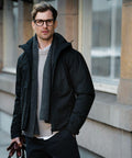 A man wearing glasses, a black jacket, a gray scarf, and holding Nimbus NB88M stands in front of a building.