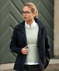 A woman with glasses and a ponytail is wearing a light green Nimbus NB89F over a white shirt, along with a black jacket. She stands in front of a textured, green background.