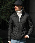 A person wearing a black quilted jacket, white turtleneck sweater, blue jeans, and a black Nimbus NB90F stands outdoors near green foliage.