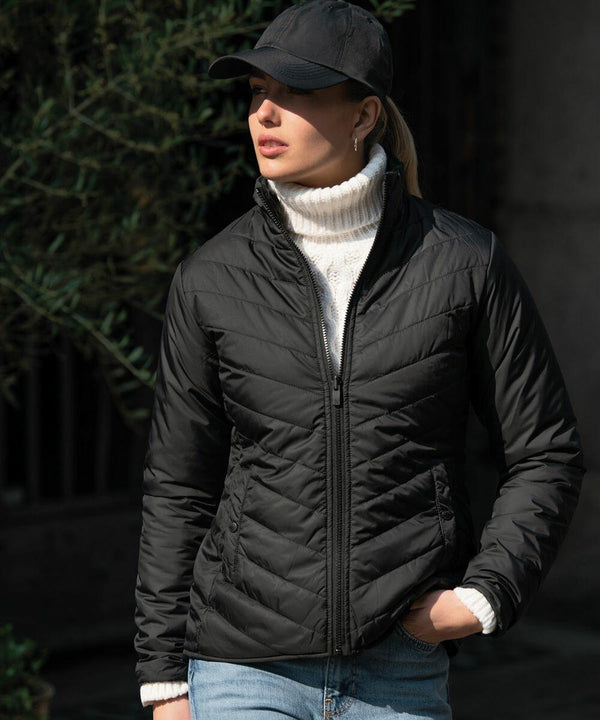 A person wearing a black quilted jacket, white turtleneck sweater, blue jeans, and a black Nimbus NB90F stands outdoors near green foliage.